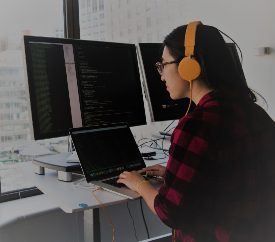 woman standing her workstation in from of her laptop and multiple computer monitor screens with head phones