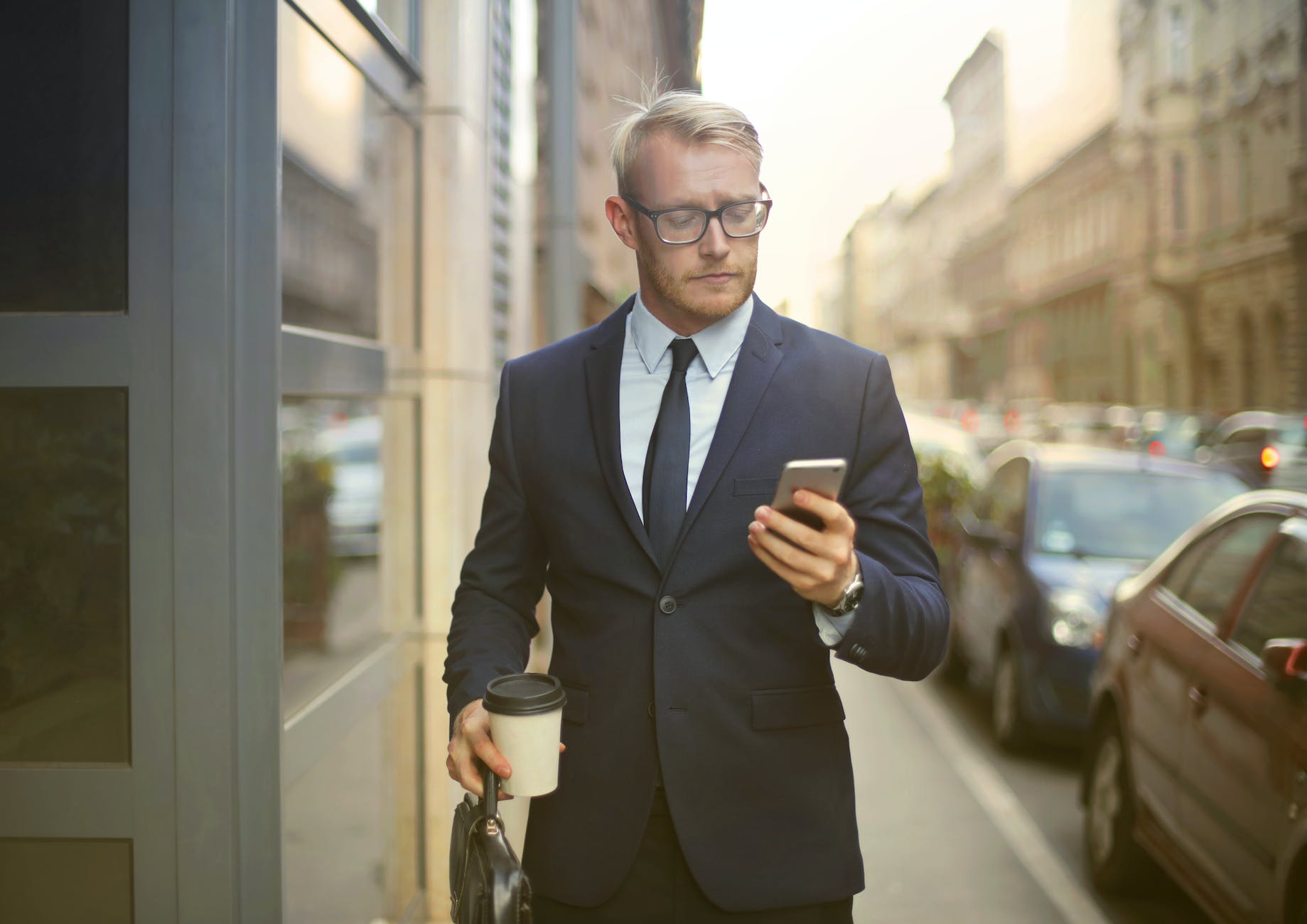 selective focus photo of walking man in black suit carrying a to go cup and briefcase while using his phone